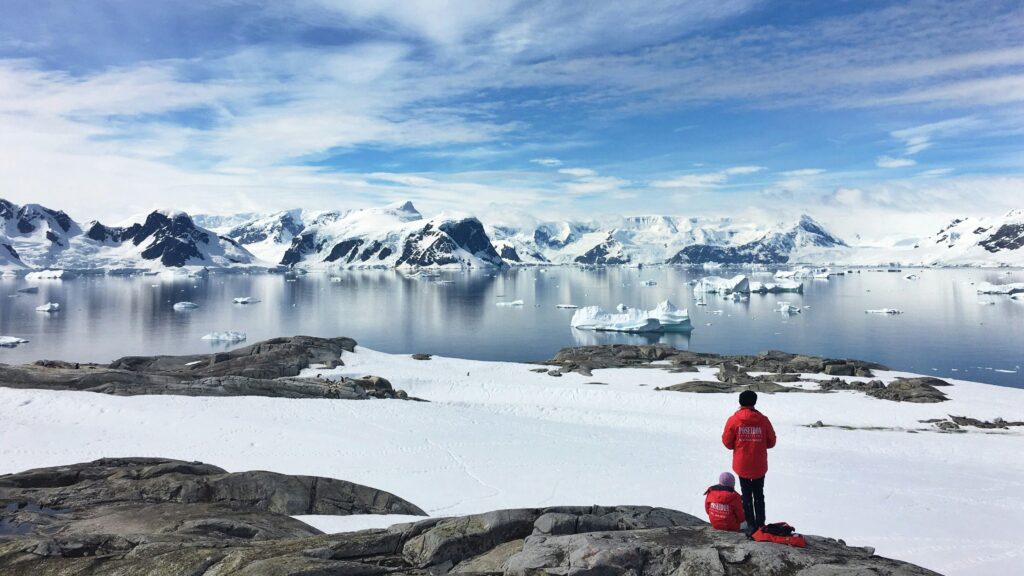 Two tourists stand in awe of the snowy landscape and majestic view offered by Antarctica.
